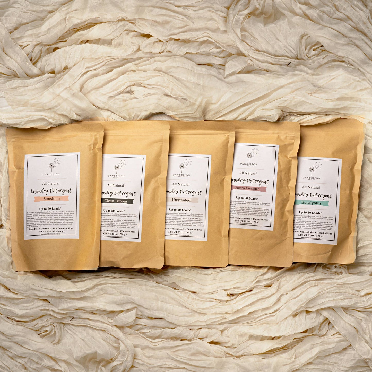 Five all-natural laundry detergent packages in kraft paper pouches with scents Sunshine, Clean Hippie, Unscented, French Lavender, and Eucalyptus on beige fabric