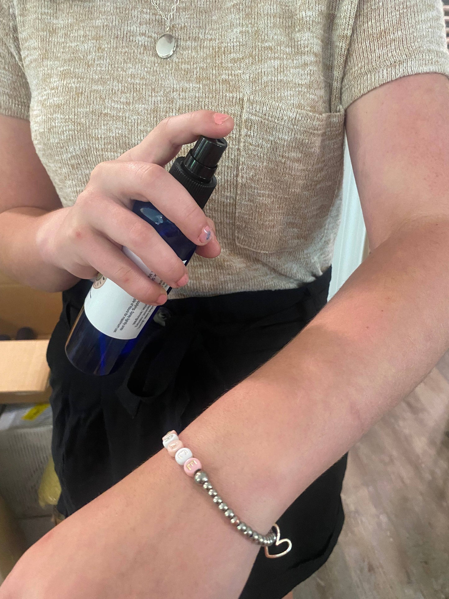 Person holding blue spray bottle applying natural skin spray on arm, wearing beige shirt and bracelet