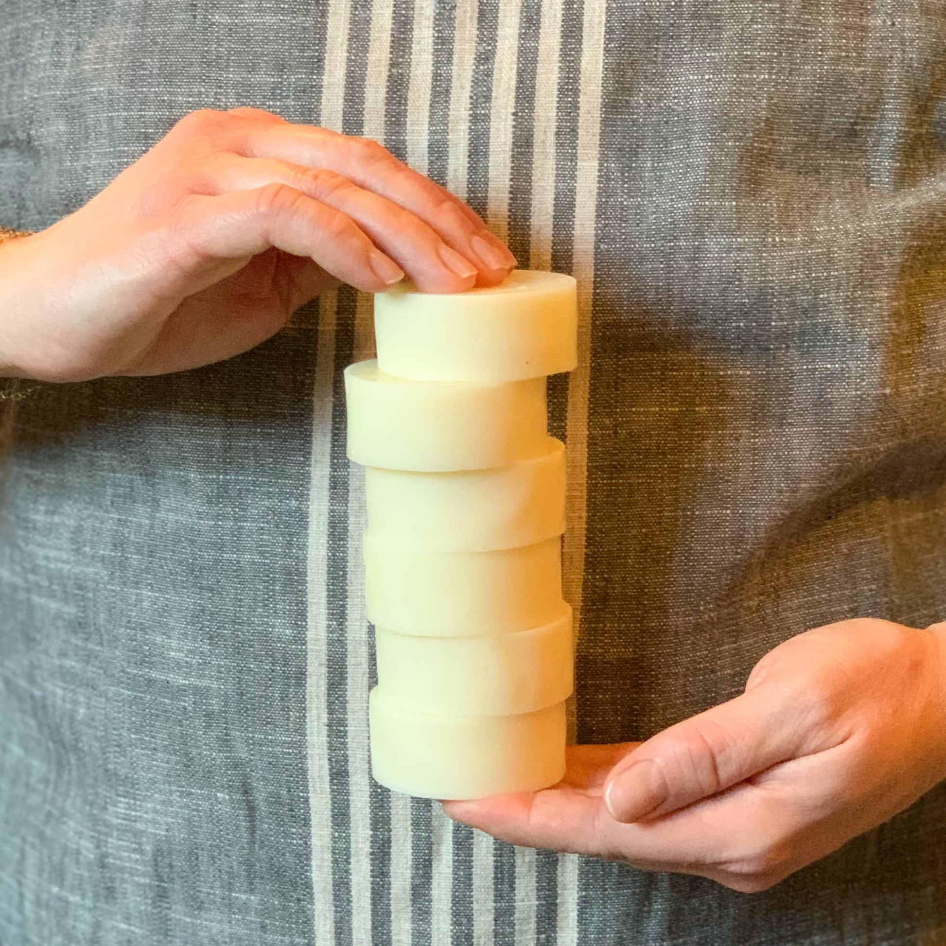 Hands stacking six round natural remedy soap bars on grey fabric background
