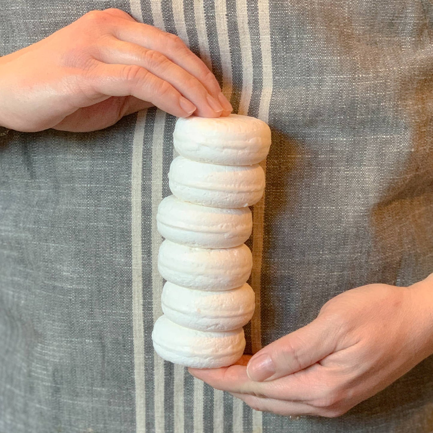 Hands holding a stack of six white round natural loofah soap bars against striped gray fabric background