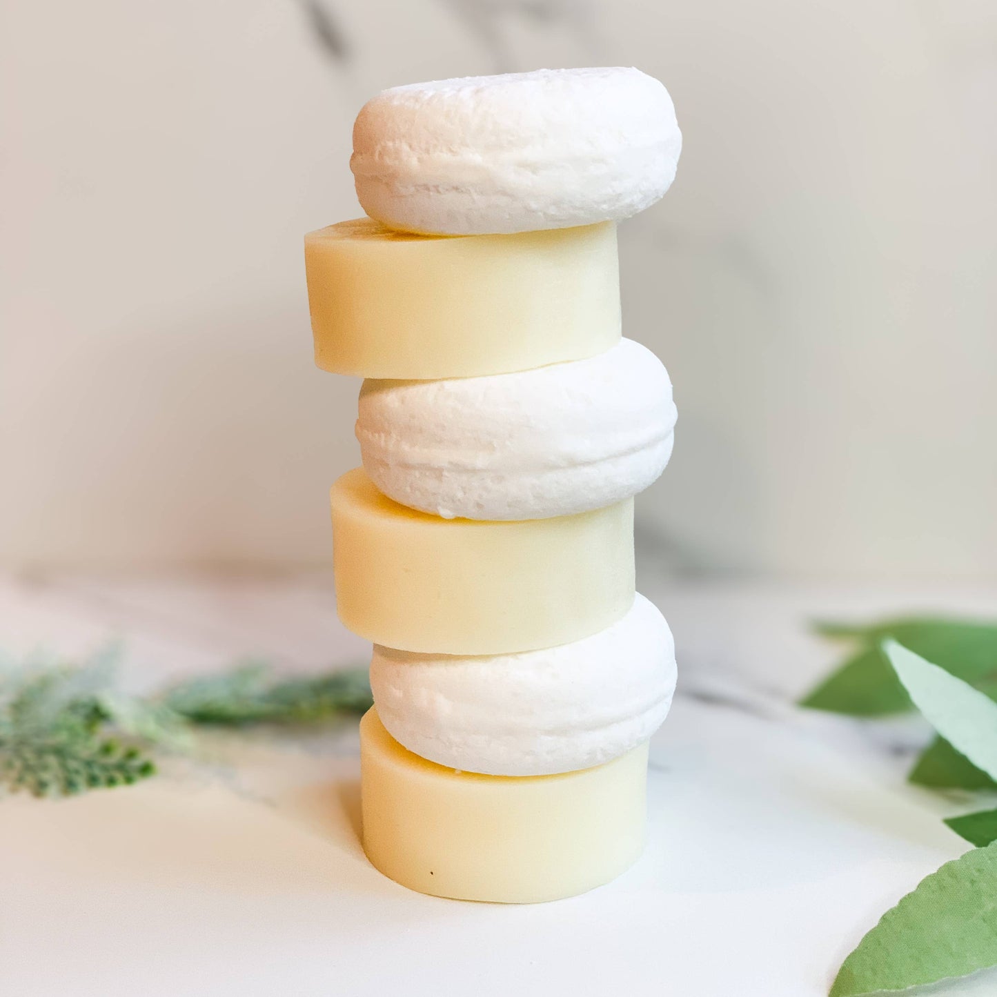 Stack of natural round solid shampoo and conditioner bars on white surface with greenery