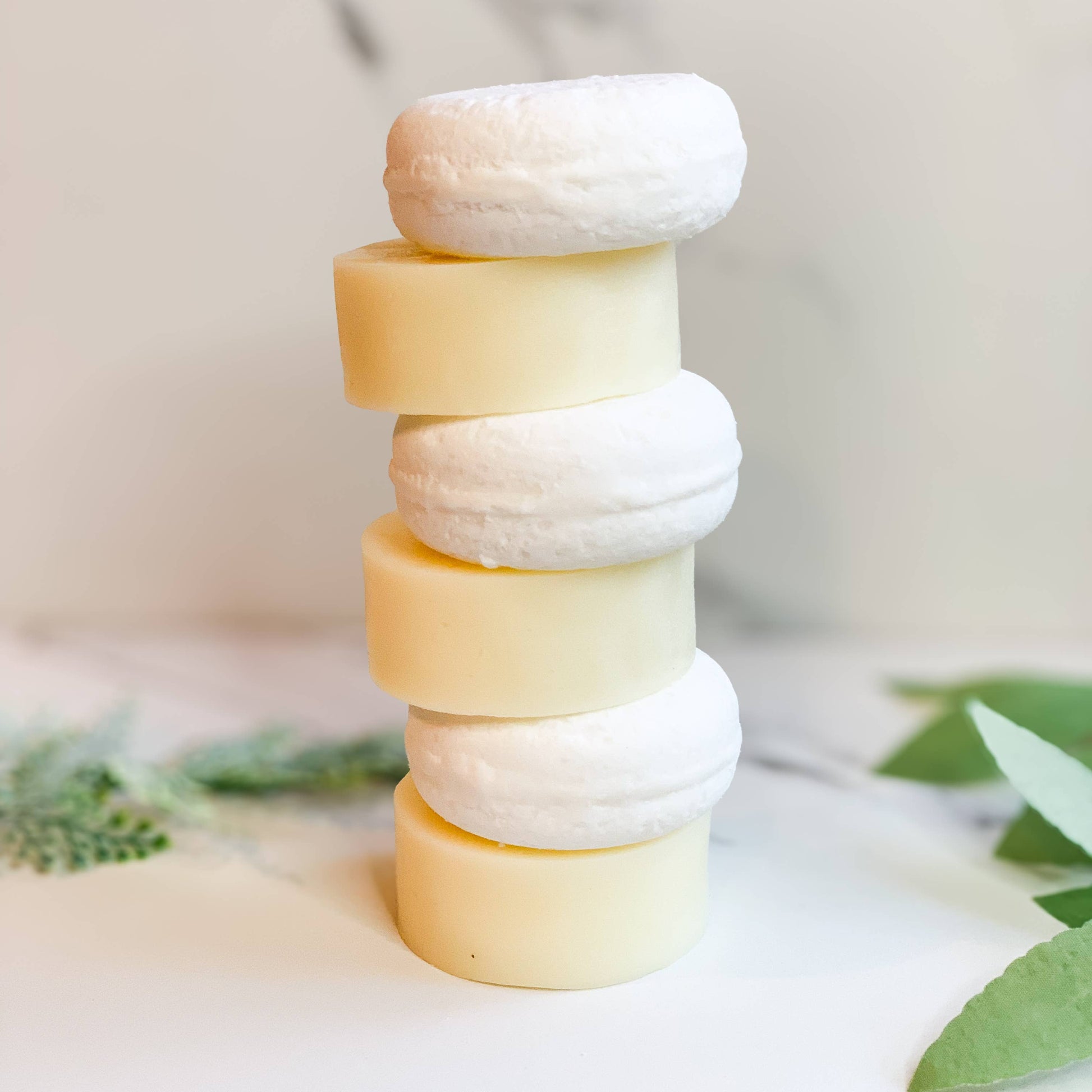 Stack of natural round solid shampoo and conditioner bars on white surface with greenery