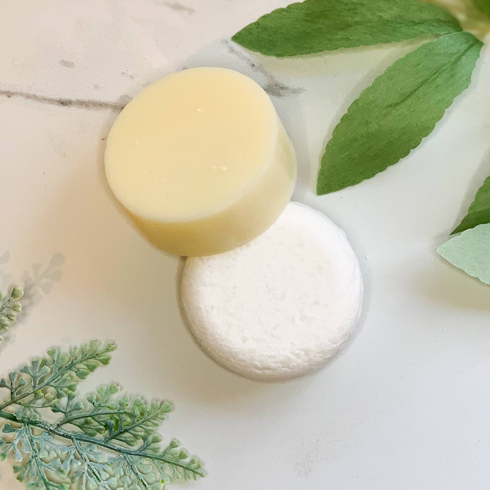 Two round natural remedy soaps, one beige and one white, on a white surface with green leaves