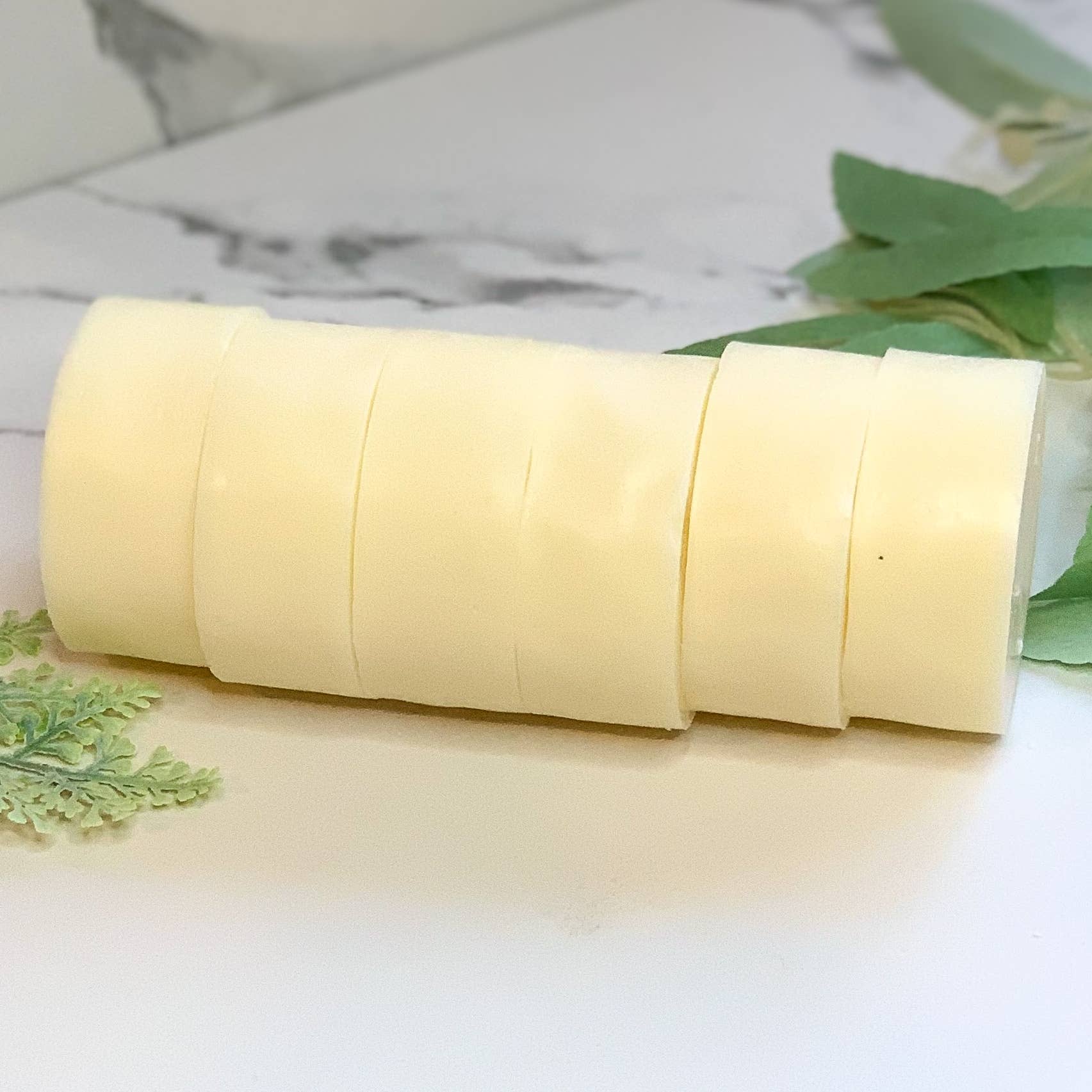 Six light yellow natural soap bars stacked horizontally on white surface with green leaves nearby