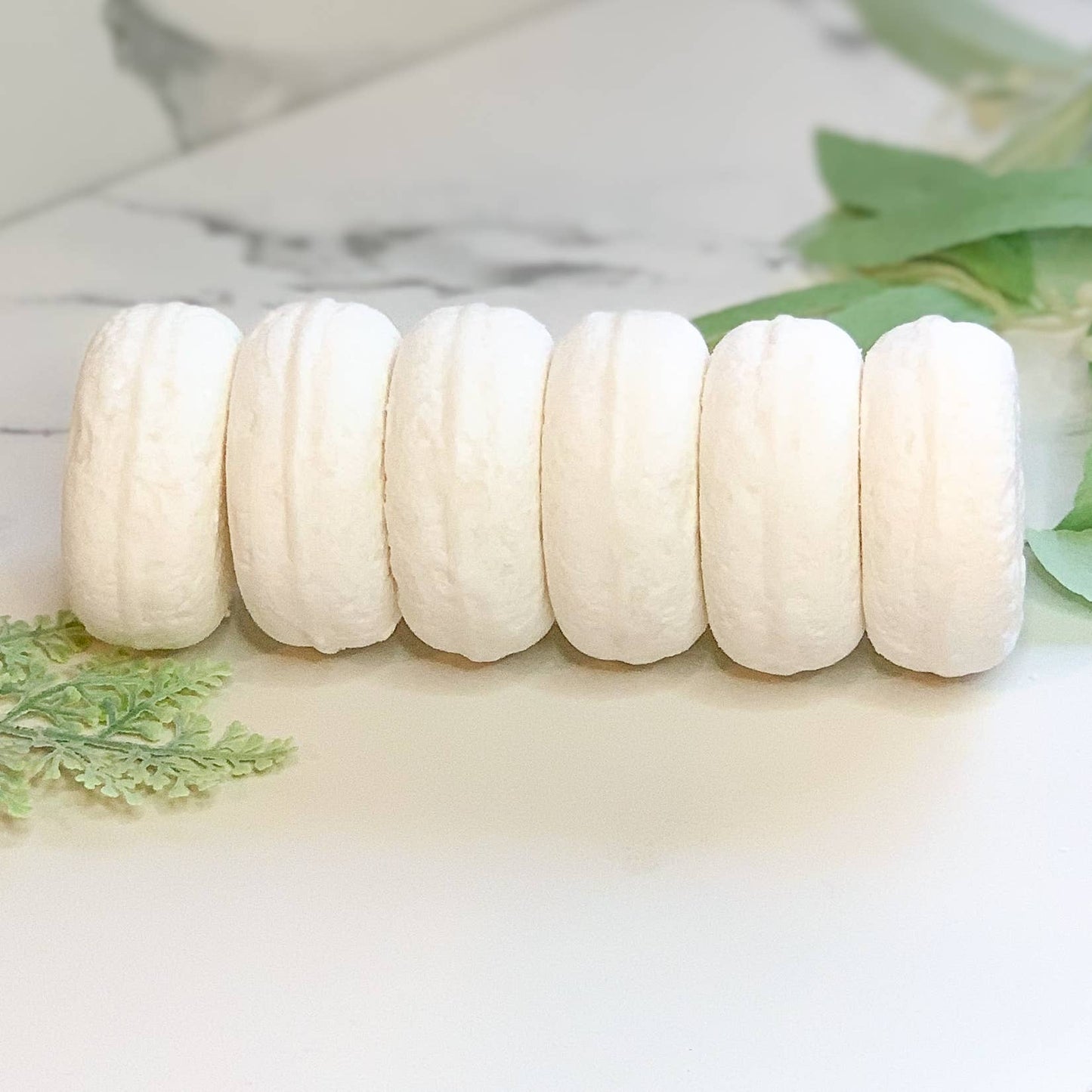Six white natural bath bombs lined up on marble surface with green leaves, Rooted In Faith Apothecary