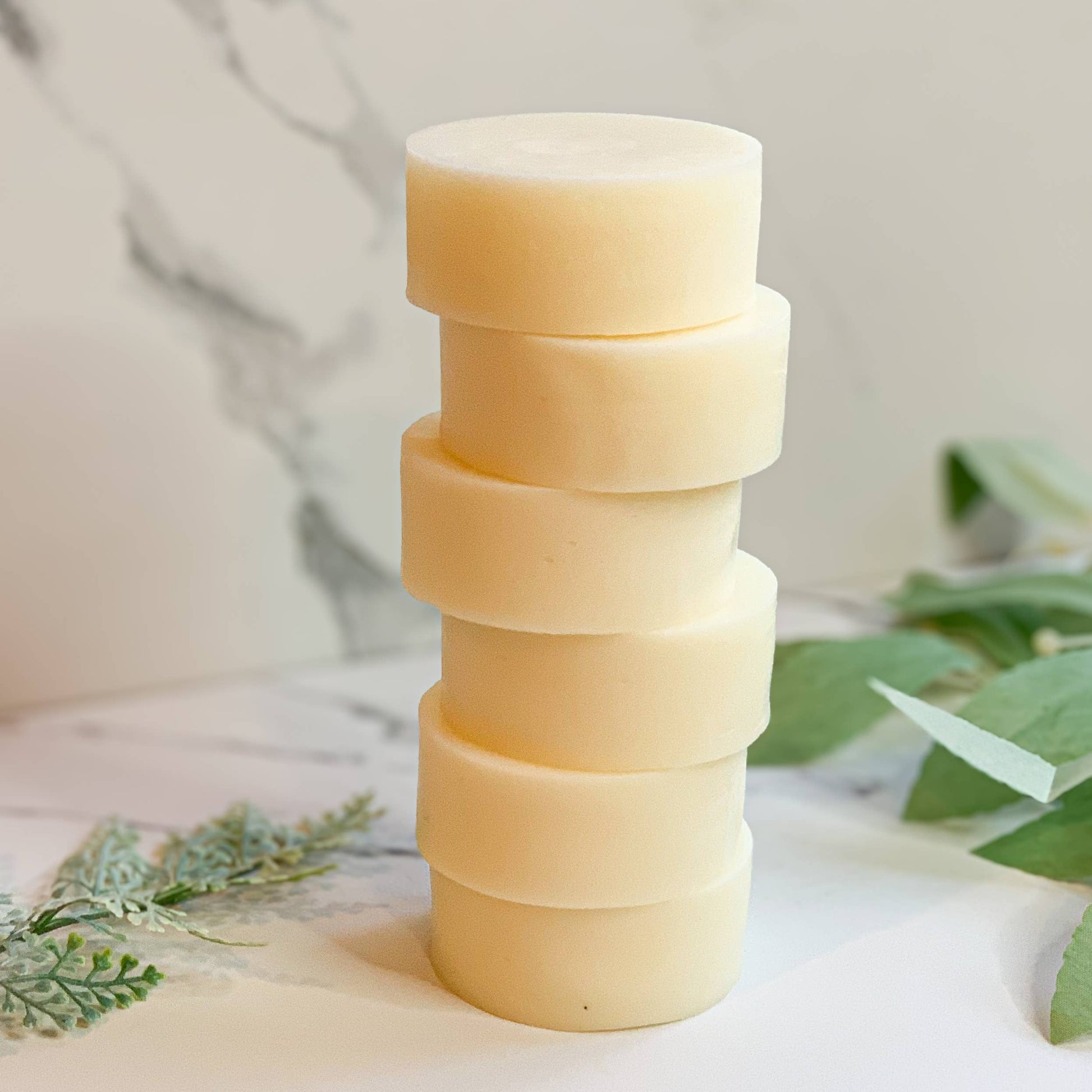 Stack of six natural beige round soap bars on marble surface with green leaves, Rooted In Faith Apothecary