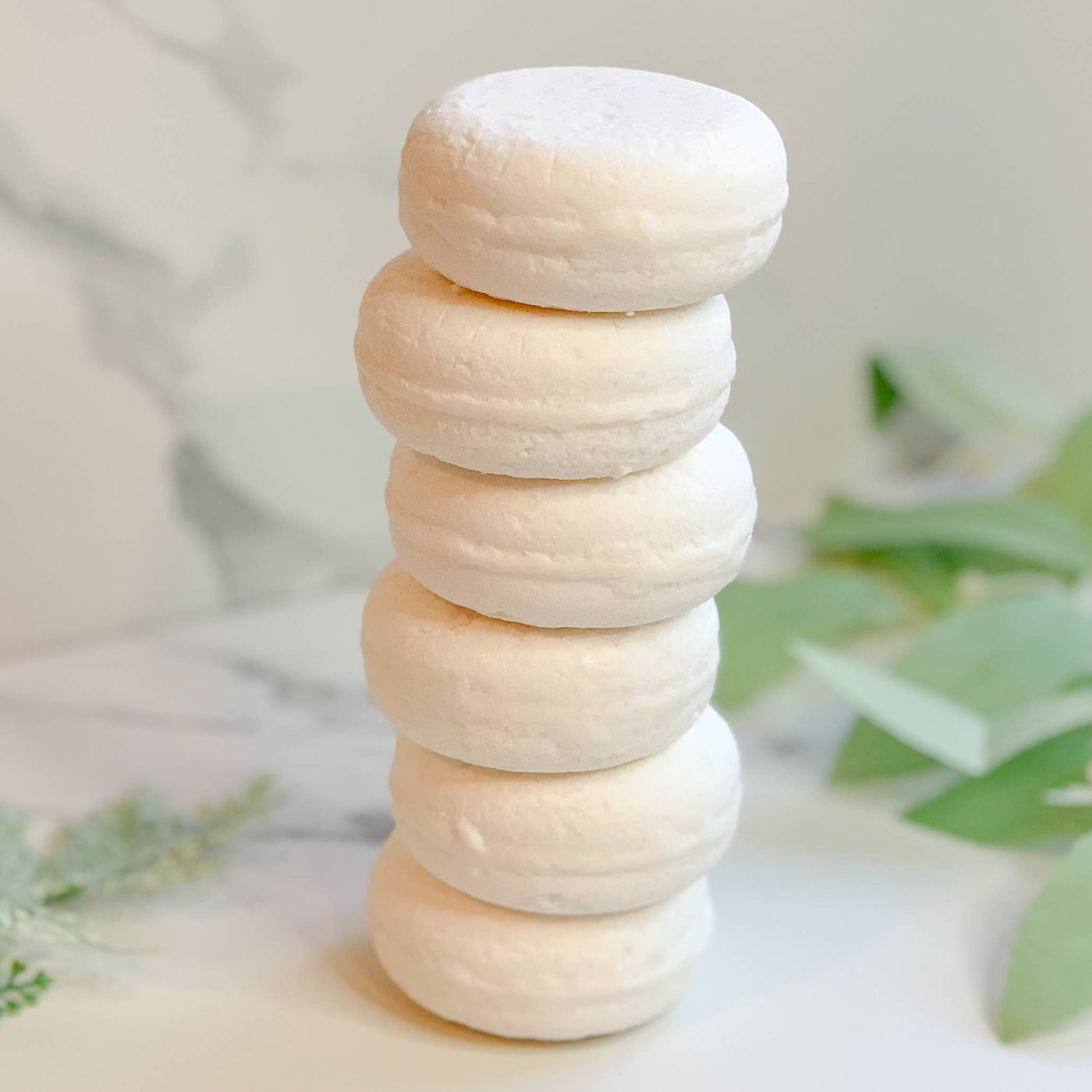 Stack of five round white natural bath bombs on a marble surface with green leaves in the background